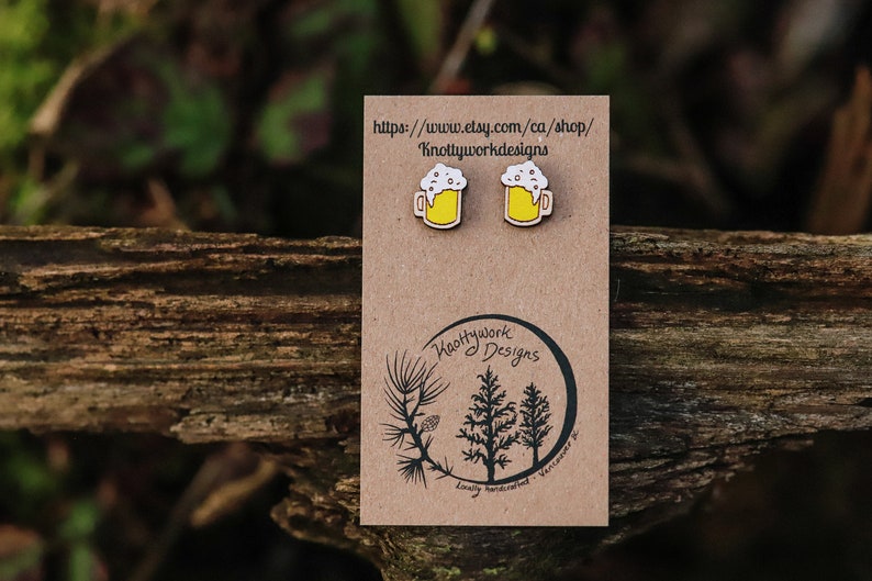 May include: A pair of wooden beer mug earrings with a white foam top and yellow liquid. The earrings are on a brown card with the text "Knottywork Designs" and a website address.