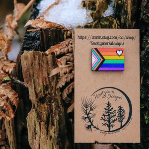 May include: A wooden pin with a rainbow flag design and a white heart. The pin is on a brown card with the text "Knottywork Designs" and the website address "https://www.etsy.com/ca/shop/Knottyworkdesigns".