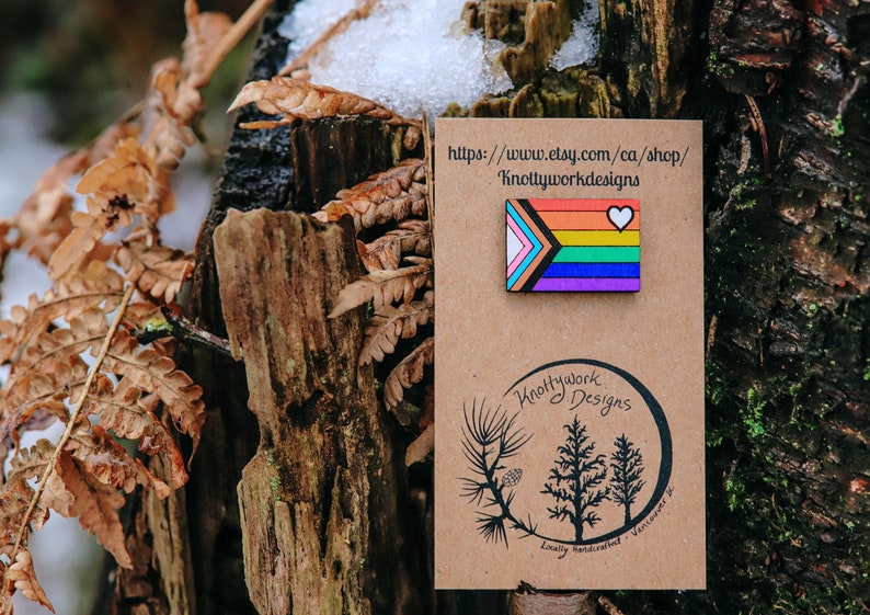 May include: A wooden pin with a rainbow flag design and a white heart. The pin is on a brown card with the text "Knottywork Designs" and the website address "https://www.etsy.com/ca/shop/Knottyworkdesigns".