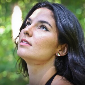 Pu&ograve; includere: Una donna con lunghi capelli scuri guarda in alto con un'espressione di speranza. Indossa una canotta nera e ha un piccolo orecchino arcobaleno all'orecchio destro.
