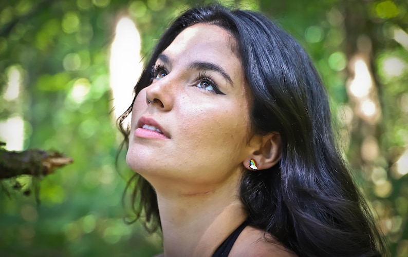 Pu&ograve; includere: Una donna con lunghi capelli scuri guarda in alto con un'espressione di speranza. Indossa una canotta nera e ha un piccolo orecchino arcobaleno all'orecchio destro.
