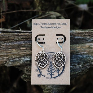 May include: A pair of wooden earrings shaped like a hop leaf with silver hooks. The earrings are displayed on a brown card with the text "https://www.etsy.com/ca/shop/KnottyWorkDesigns".