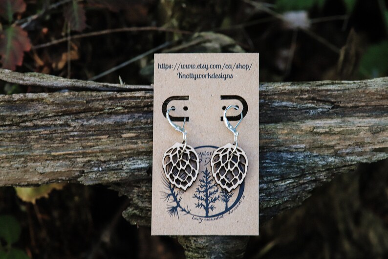 May include: A pair of wooden earrings shaped like a hop leaf with silver hooks. The earrings are displayed on a brown card with the text "https://www.etsy.com/ca/shop/KnottyWorkDesigns".