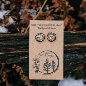 May include: A pair of wooden earrings with a white and brown floral design. The earrings are displayed on a brown card with the text "Knottywork Designs" and a website address.