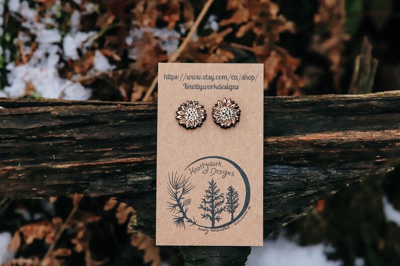 May include: A pair of wooden earrings with a white and brown floral design. The earrings are displayed on a brown card with the text "Knottywork Designs" and a website address.