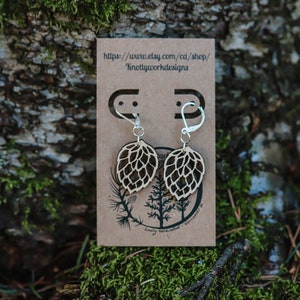 May include: A pair of wooden earrings shaped like a hop cone, with silver hooks. The earrings are displayed on a brown card with the text "https://www.etsy.com/ca/shop/Knottywooddesigns".