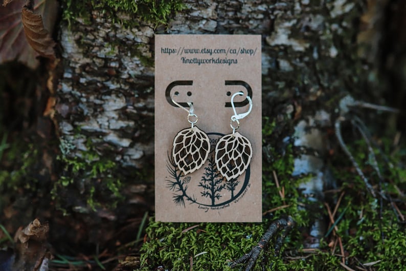 May include: A pair of wooden earrings shaped like a hop cone, with silver hooks. The earrings are displayed on a brown card with the text "https://www.etsy.com/ca/shop/Knottywooddesigns".