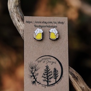 May include: A pair of wooden beer mug earrings with yellow mugs and white foam. The earrings are on a brown card with the text "https://www.etsy.com/ca/shop/Knottyworkdesigns" and a logo that says "Knottywork Designs" with a tree design.