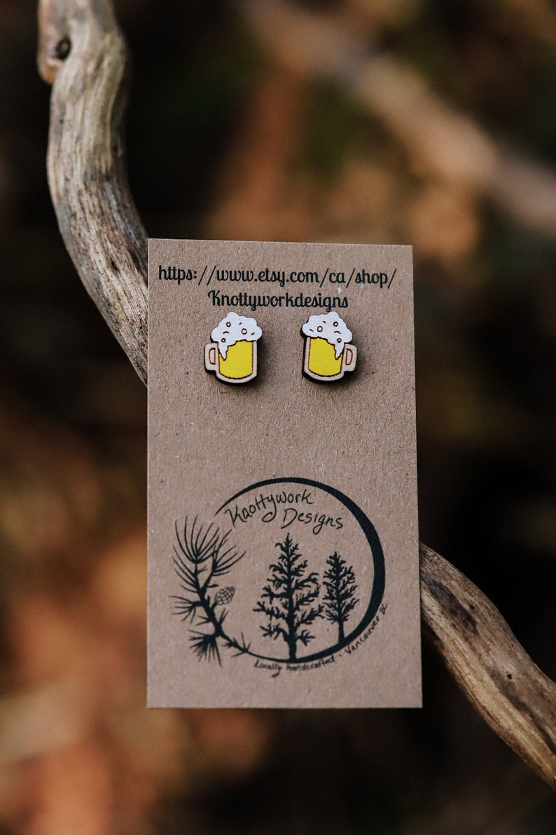 May include: A pair of wooden beer mug earrings with yellow mugs and white foam. The earrings are on a brown card with the text "https://www.etsy.com/ca/shop/Knottyworkdesigns" and a logo that says "Knottywork Designs" with a tree design.