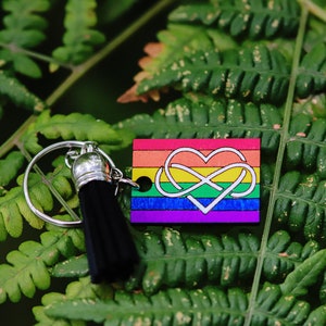 Könnte beinhalten: Ein regenbogenfarbener Holzschlüsselanhänger mit einem weißen Unendlichkeitszeichen in einem Herzen. Der Schlüsselanhänger hat einen schwarzen Quasten.