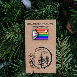 Puede incluir: Un pin de madera con un diseño de bandera arcoíris y un pequeño corazón blanco. El pin está en una tarjeta marrón con el texto "Knottywork Designs" y una dirección de sitio web: "https://www.etsy.com/ca/shop/Knottyworkdesigns".