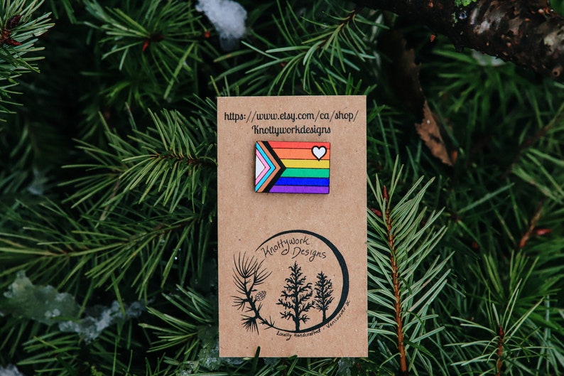 May include: A wooden pin with a rainbow flag design and a small white heart. The pin is on a brown card with the text "Knottywork Designs" and a website address: "https://www.etsy.com/ca/shop/Knottyworkdesigns".