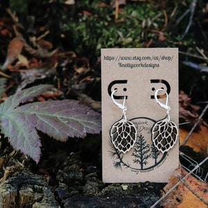 May include: A pair of wooden earrings shaped like a hop cone, with silver hooks. The earrings are displayed on a brown card with the text "https://www.etsy.com/ca/shop/KnottyworkDesigns".