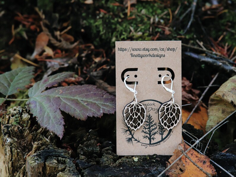 May include: A pair of wooden earrings shaped like a hop cone, with silver hooks. The earrings are displayed on a brown card with the text "https://www.etsy.com/ca/shop/KnottyworkDesigns".