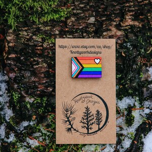 May include: A wooden pin with a rainbow flag design and a white heart. The pin is on a brown card with the text "Knottywork Designs" and a website address. The card is on a background of green moss and snow.