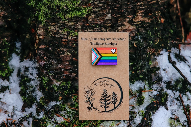 May include: A wooden pin with a rainbow flag design and a white heart. The pin is on a brown card with the text "Knottywork Designs" and a website address. The card is on a background of green moss and snow.