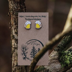 May include: A pair of wooden beer mug earrings with yellow beer and white foam. The earrings are on a brown card with the text "Knottywork Designs" and a website address.