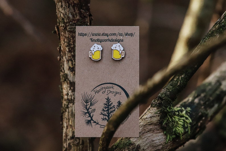 May include: A pair of wooden beer mug earrings with yellow beer and white foam. The earrings are on a brown card with the text "Knottywork Designs" and a website address.