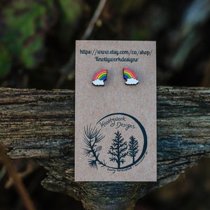 Pu&ograve; includere: Un paio di orecchini in legno a forma di arcobaleno e nuvola su una carta marrone con il testo "Knottywork Designs" e un indirizzo di sito web.