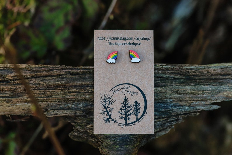 Pu&ograve; includere: Un paio di orecchini in legno a forma di arcobaleno e nuvola su una carta marrone con il testo "Knottywork Designs" e un indirizzo di sito web.