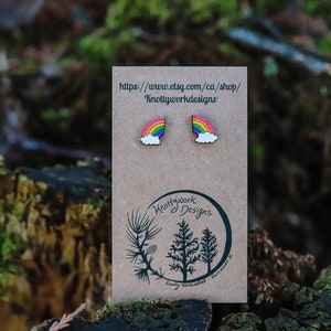 Pu&ograve; includere: Un paio di orecchini in legno arcobaleno con un design a nuvola. Gli orecchini sono esposti su una carta marrone con il testo "Knottywork Designs" e un indirizzo di sito web.