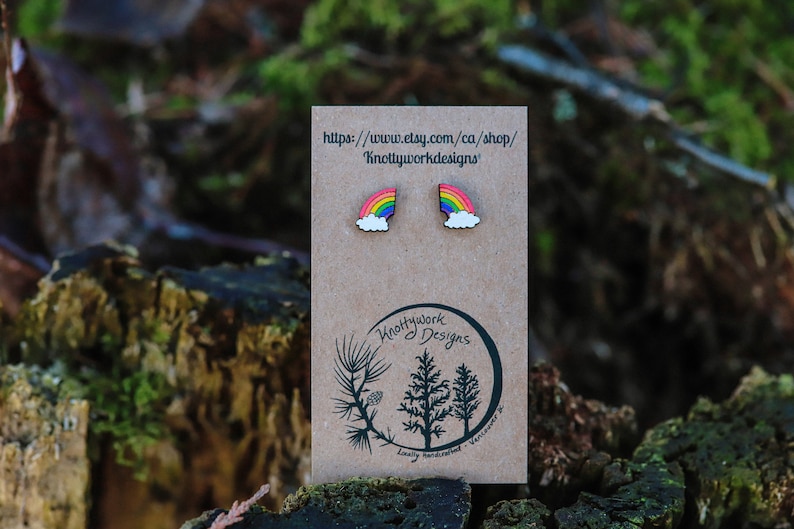 Pu&ograve; includere: Un paio di orecchini in legno arcobaleno con un design a nuvola. Gli orecchini sono esposti su una carta marrone con il testo "Knottywork Designs" e un indirizzo di sito web.