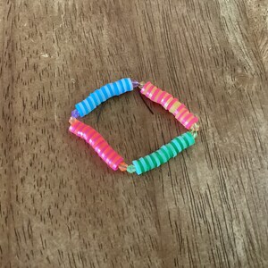 Puede incluir: Un brazalete colorido hecho de cuentas de plástico rayadas. El brazalete está hecho de cuatro colores diferentes: azul, rosa, verde y amarillo.
