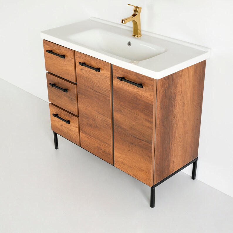 Puede incluir: Un tocador de ba&ntilde;o de madera marr&oacute;n con un lavabo de porcelana blanca y herrajes de metal negro. El tocador tiene tres cajones y dos puertas. El tocador est&aacute; sobre patas de metal negro.