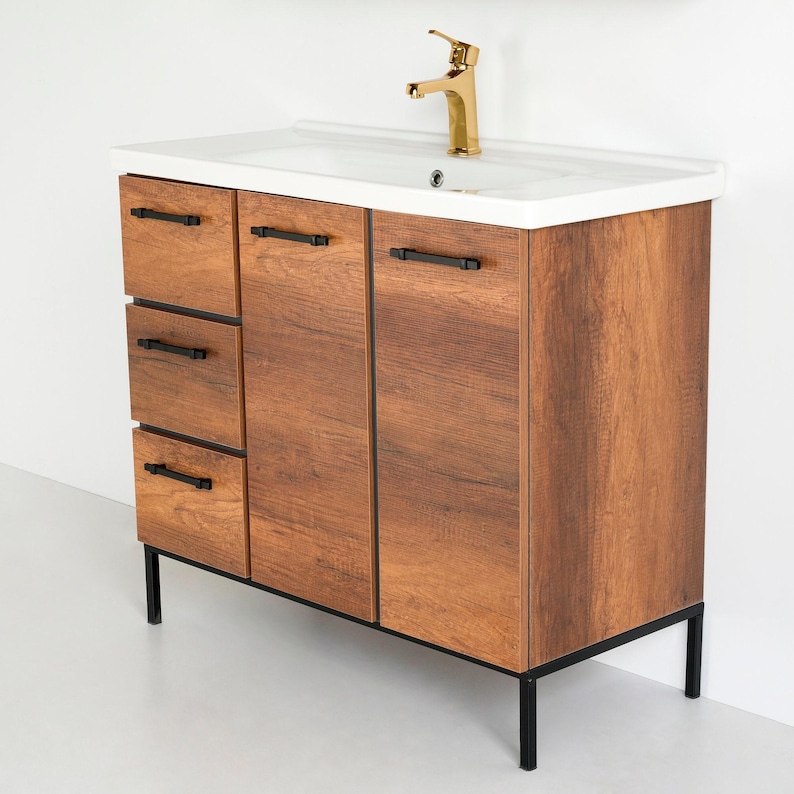 May include: A brown wooden bathroom vanity with black metal legs and black hardware. The vanity has three drawers and two doors. The vanity top is white and has a single faucet.