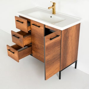 Puede incluir: Un tocador de ba&ntilde;o de madera marr&oacute;n con un lavabo de porcelana blanca y herrajes negros. El tocador tiene tres cajones y dos puertas. Las puertas est&aacute;n abiertas y los cajones est&aacute;n sacados. El tocador tiene patas de metal negro.