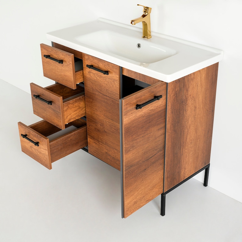 Puede incluir: Un tocador de ba&ntilde;o de madera marr&oacute;n con un lavabo de porcelana blanca y herrajes negros. El tocador tiene tres cajones y dos puertas. Las puertas est&aacute;n abiertas y los cajones est&aacute;n sacados. El tocador tiene patas de metal negro.