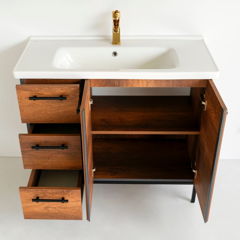 Puede incluir: Un tocador de ba&ntilde;o de madera marr&oacute;n con un lavabo de porcelana blanca y un grifo dorado. El tocador tiene tres cajones y dos puertas que se abren para revelar estantes. El tocador est&aacute; sobre un fondo blanco.