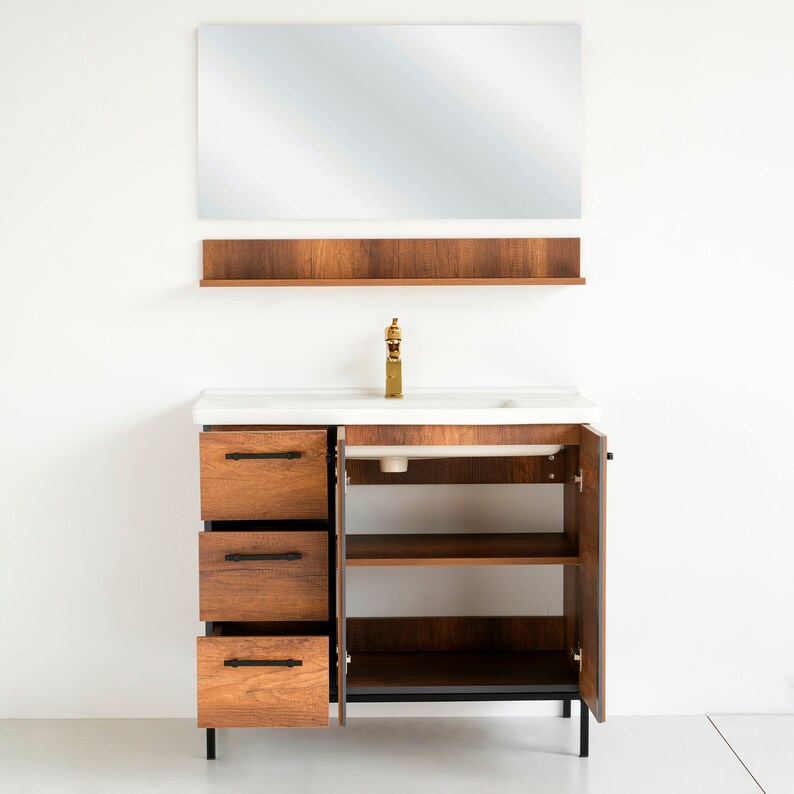 Puede incluir: Un tocador de ba&ntilde;o de madera con un lavabo de porcelana blanca y un grifo dorado. El tocador tiene tres cajones y dos estantes abiertos. Un espejo con una repisa de madera est&aacute; montado sobre el tocador.