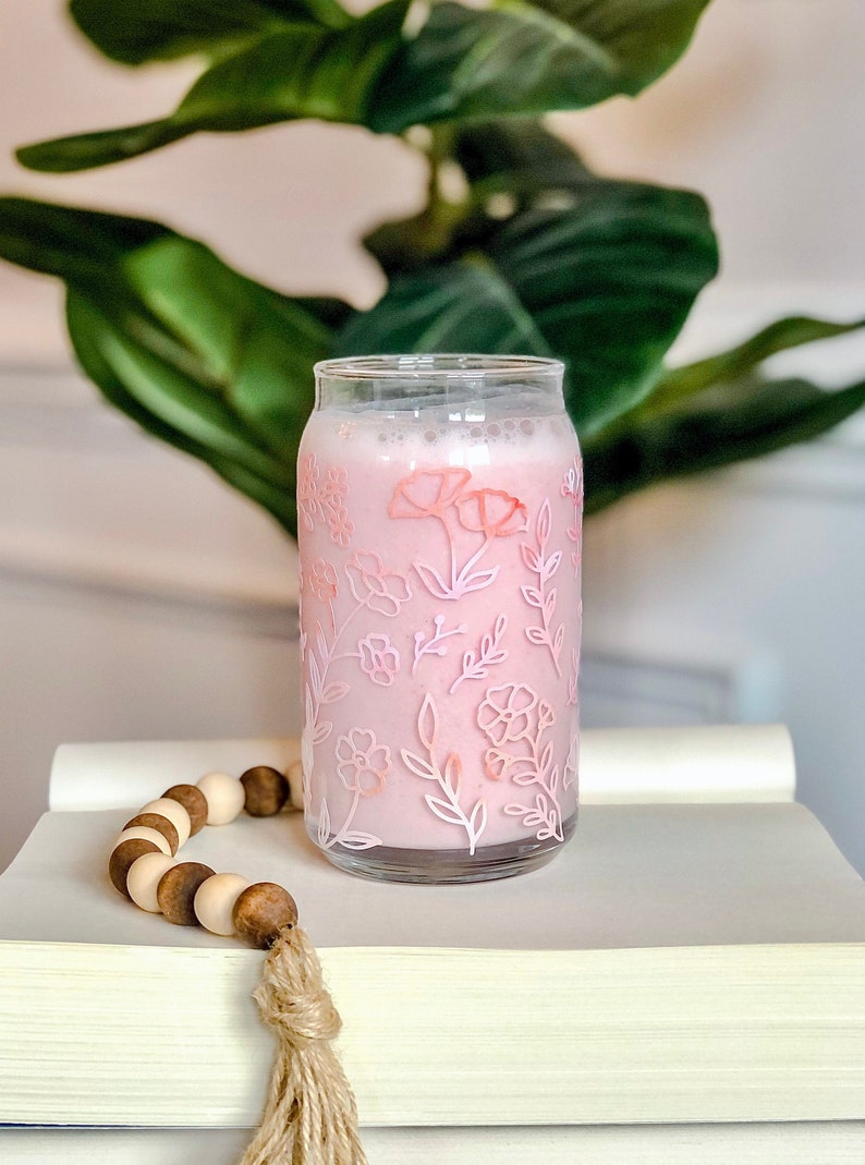 May include: A glass with a pink floral design filled with a pink beverage. The glass is sitting on a stack of books with a wooden bead garland.