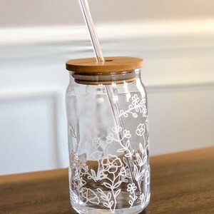 May include: A clear glass drinking jar with a white floral design and a bamboo lid. A clear glass straw is inserted through the lid.