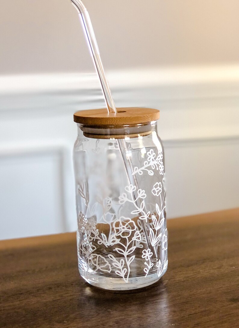May include: A clear glass drinking jar with a white floral design and a bamboo lid. A clear glass straw is inserted through the lid.