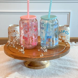 May include: Two glass tumblers with lids and straws. The tumblers are decorated with cherries and bows. One tumbler is pink with red cherries and the other is blue with blue cherries. The tumblers are on a wooden cake stand.