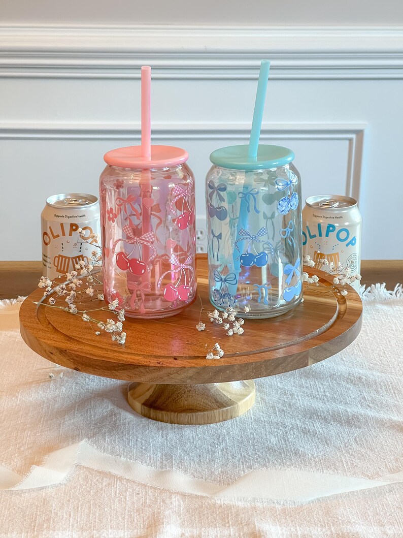 May include: Two glass tumblers with lids and straws. The tumblers are decorated with cherries and bows. One tumbler is pink with red cherries and the other is blue with blue cherries. The tumblers are on a wooden cake stand.
