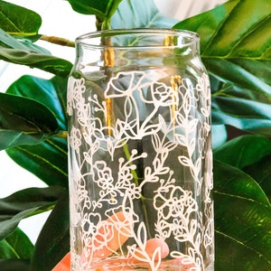 May include: A clear glass jar with a white floral design painted on the outside. The jar is being held by a hand.