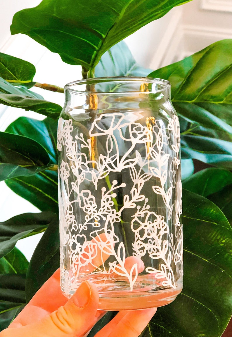 May include: A clear glass jar with a white floral design painted on the outside. The jar is being held by a hand.