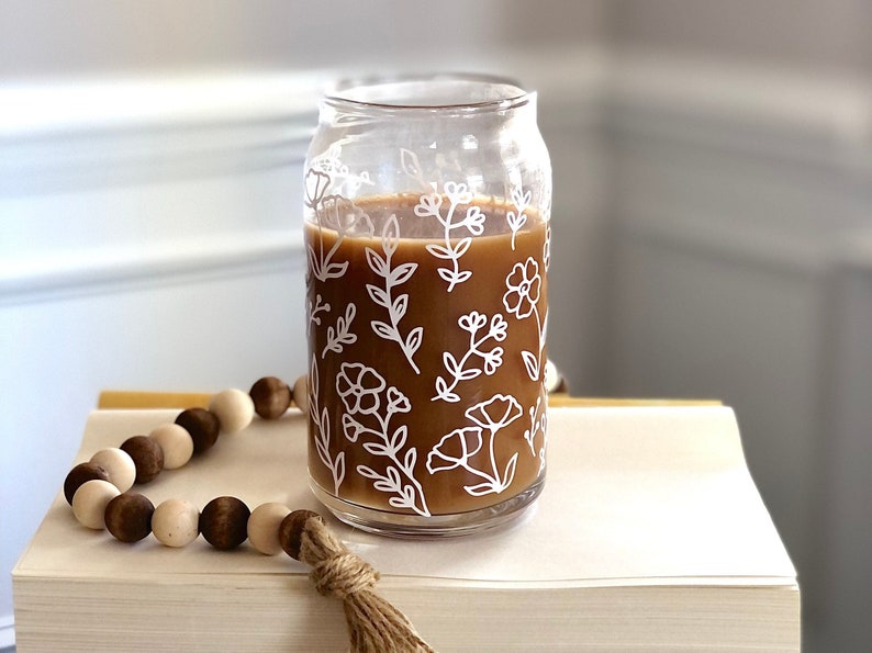 May include: A glass mug with a floral design and brown liquid inside. The mug is sitting on a brown surface with a wooden bead garland.
