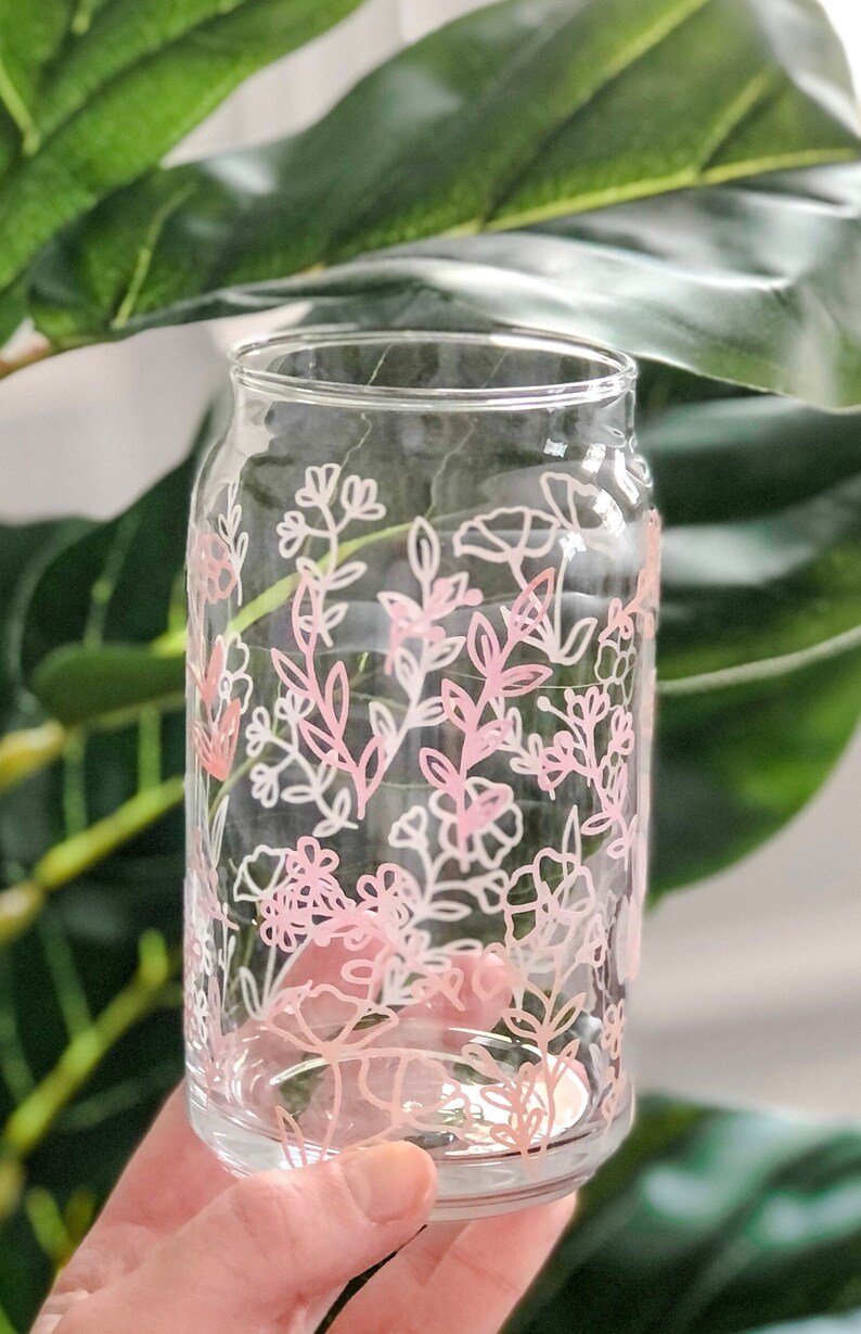 May include: A clear glass drinking glass with a pink and white floral design. The design features delicate flowers and leaves.