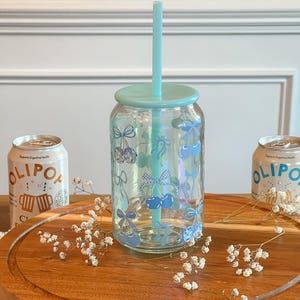 May include: A clear glass reusable can with a blue lid and a blue straw. The can has a blue and white checkered pattern with blue bows and cherries. The can is sitting on a wooden surface with white flowers.
