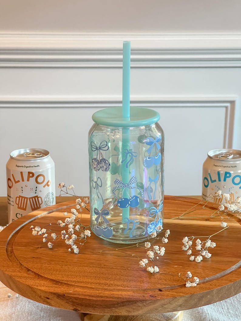 May include: A clear glass reusable can with a blue lid and a blue straw. The can has a blue and white checkered pattern with blue bows and cherries. The can is sitting on a wooden surface with white flowers.