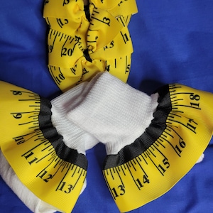 May include: A pair of white socks with yellow ruffled cuffs featuring a black measuring tape print with inch markings, accompanied by a matching yellow hair bow with a black measuring tape print and black centre knot on a brown card backing.