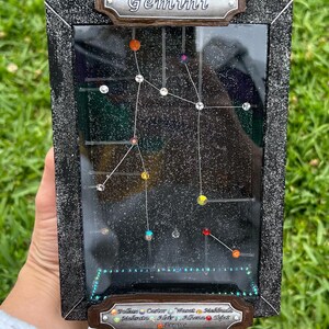 Puede incluir: Una obra de arte enmarcada que representa la constelación de Géminis. El diseño presenta un fondo negro con un patrón de constelación, utilizando cuentas de colores para representar las estrellas. El marco oscuro tiene un acabado texturizado, y la palabra "Géminis" está en la parte superior.