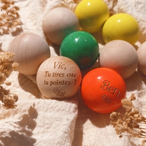 May include: A collection of wooden and plastic balls with personalized messages. The balls are in various colors, including yellow, green, orange, and natural wood. The messages on the balls include "Vic, Tu tires ou tu pointes!" and "Ben".