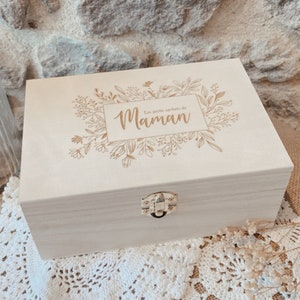 Peut inclure: Une boîte en bois de couleur claire avec un motif floral et le texte "Les petits secrets de Maman" en or. La boîte a un fermoir en métal et est posée sur un napperon en dentelle blanche.