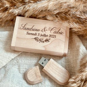 Peut inclure: Une clé USB en bois et son coffret assorti, gravés des noms "Sandrine & Cédric" et de la date "Samedi 5 juillet 2025". La clé USB est partiellement ouverte, révélant le connecteur métallique. L'ensemble repose sur une surface texturée blanc cassé.
