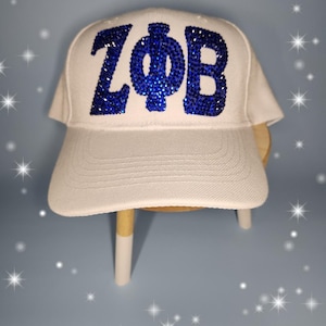 Puede incluir: Gorra de b&eacute;isbol blanca con letras de pedrer&iacute;a azul que dicen "ZФB".